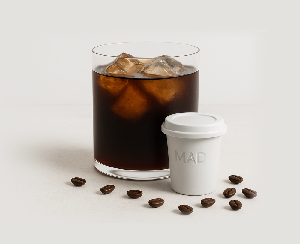 Glass of iced coffee with a white cup labeled 'MAD' and scattered coffee beans on a light background